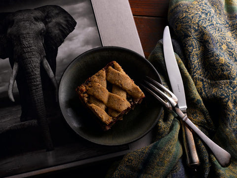 A raspberry hazelnut bar on a dark green plate with a fork and knife beside it, placed on a wooden table with a patterned cloth and a black and white photo of an elephant.