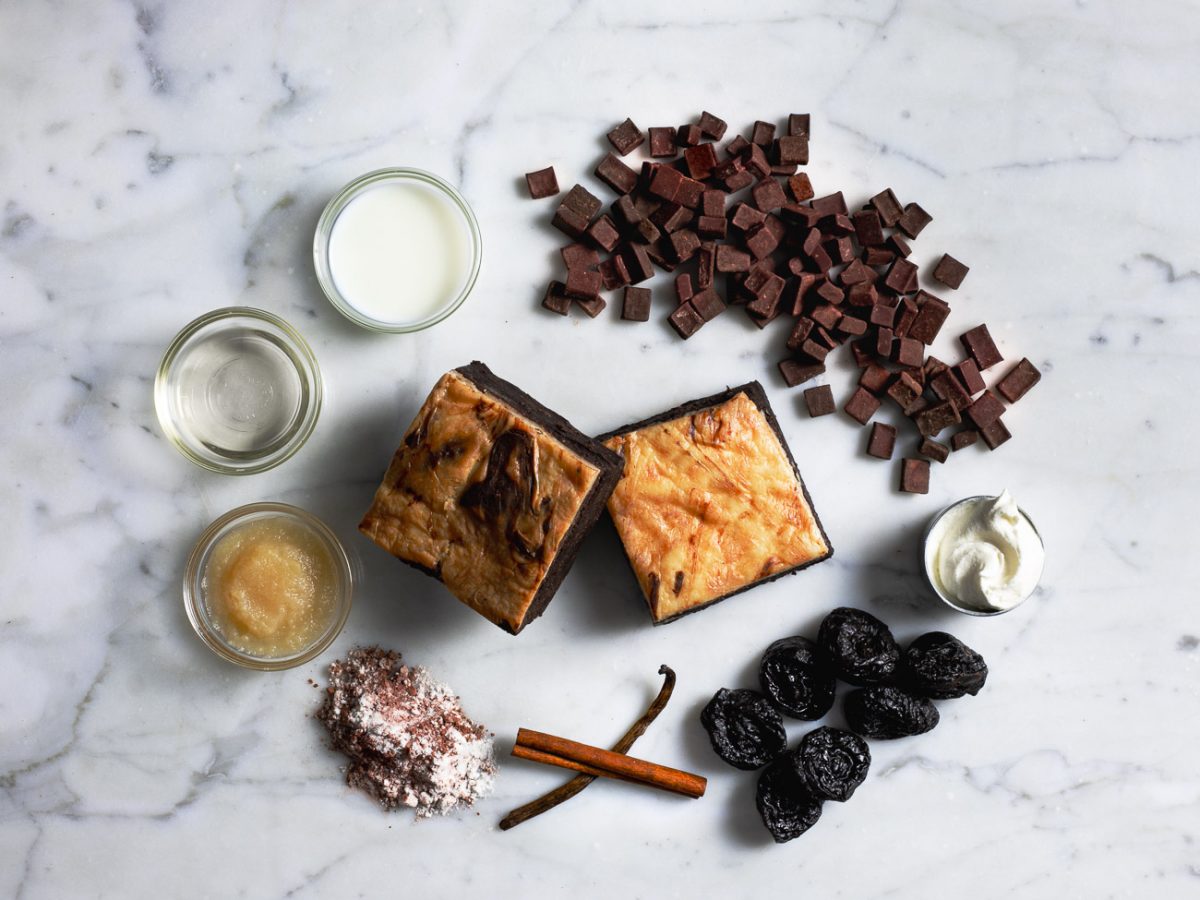 Ingredients for 2 cheescake brownies including chocolate chunks, prunes, cinnamon sticks, vanilla bean, applesauce, flour, milk, and cream cheese on a marble surface.