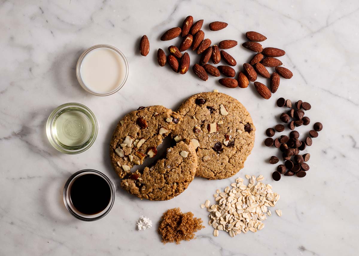 Top view of two salted almond chocolate chip cookies surrounded by ingredients: almonds, chocolate chips, oat flour, sugar, oil, vanilla, and milk on a marble surface.